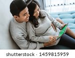 happiness of couples holding the wife's pregnant stomach while using digital tablet, sitting on the couch beside the ornamental plants