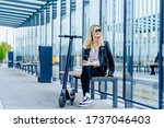 Young millennial woman relaxing after rides a cool electric scooter past, sitting at bus stop on a sunny day. Smiling Blond fashionable girl in black clothes while waiting her tram on empty tram stop.