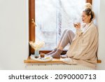 Happy blond woman with bunch hairstyle warming and cover knitted plaid enjoying in her coffee time by the window in cold winter day. Peace of mind and mental health.