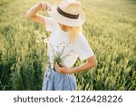 Small photo of Gorgeous and creative caucasian woman decorate dress from chamomile cinching with belt around waste outfit, standing in wheat field at summer time in sunset. Woman unity nature concept.