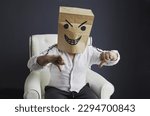 A man with a paper bag on his head, with an evil smiley face drawn, sits on a white chair, gloats emotionally. Emotions and gestures.