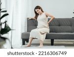 Small photo of Full body of a beautiful pregnant asian woman is sitting on a couch in the living room, having a back pain as she carries the baby in her tummy. Pregnant problem.