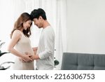 Small photo of A happy asian couple. A beautiful pregnant wife and her handsome husband are smiling happily with their heads touched and their hands grabbing the wife's tummy to feel their baby.