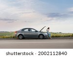 A frustrated young girl stands near a broken-down car in the middle of the highway during sunset. Breakdown and repair of the car. Waiting for help.