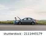 A young girl stands near a broken car in the middle of the highway during sunset and tries to call for help on the phone and start the car. Waiting for help. Car service. Car breakdown on the road.
