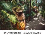 Young beautiful woman stands in the shade of palm trees, view from the back. A girl walks among tropical greenery on a bright sunny day in Mexico, Tulum 