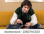 Small photo of A boy or young man sitting on a couch or sofa and playing video games with a controller while wearing headphones. new player. noob gamer.