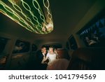 Beautiful wedding photosession. Handsome groom in blue formal suit and bow tie with boutonniere kisses hand of the elegant bride in white dress and veil with a beautiful hairdress in a limousine.