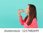 Side view of young woman in knitted pink sweater with closed eyes hold in hand, eating eclair cake isolated on blue turquoise background, studio portrait. People lifestyle concept. Mock up copy space