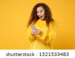 Surprised young african american girl in fur sweater posing isolated on yellow orange background, studio portrait. People lifestyle concept. Mock up copy space. Using mobile phone, typing sms message