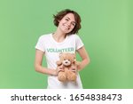 Pretty young brunette woman in white volunteer t-shirt isolated on pastel green background in studio. Voluntary free work assistance help charity grace teamwork concept. Holding teddy bear plush toy
