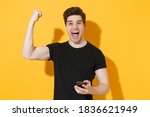 Screaming fun young man 20s in casual black t-shirt posing isolated on yellow background studio portrait. People emotions lifestyle concept. Mock up copy space. Using mobile phone doing winner gesture