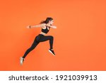 Full length side view of portrait of young fitness sporty woman 20s wearing black sportswear posing training working out jumping like running looking aside isolated on orange color background studio