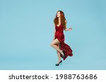 Full length of young caucasian stunning smiling readhead curly woman in red party evening bordo dress gown walk dancing fluttering dress look aside isolated on pastel blue background studio portrait.