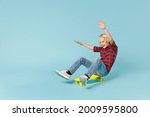 Full body length funny little curly kid boy 13s in basic red checkered shirt sitting riding on skateboard rising hands isolated on blue background children studio portrait. Childhood lifestyle concept