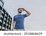 Bottom view young strong sporty athletic toned fit sportsman man 20s in blue casual t-shirt walking hold hand at forehead look far away distance in summer cloudy day morning near hotel black building