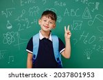 Young happy male kid school boy 5-6 years old in t-shirt backpack showing victory sign isolated on green wall chalk blackboard background studio. Childhood children kids education lifestyle concept.