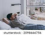 Side viw calm peaceful minded young man 20s wearing pajamas grey t-shirt lying in bed sleep slumber resting look at window relaxing at home indoors bedroom. Good mood night morning bedtime concept.