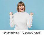 Small photo of Elderly overjoyed happy woman 50s wearing white knitted sweater doing winner gesture celebrate clenching fists say yes isolated on plain blue color background studio portrait. People lifestyle concept