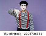 Small photo of Frowning upset distempered unnerved young mime man with white face mask wears striped shirt beret showing thumb down dislike gesture isolated on plain pastel light violet background studio portrait