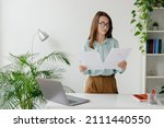 Young successful employee business woman in blue shirt hold paper account documents work stand at workplace white desk with laptop pc computer at light modern office indoors Achievement career concept