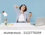 Young dreamful secretary employee business woman in casual shirt sit work at white office desk with pc laptop hold cup of coffee hold hand behind neck head isolated on pastel blue background studio.