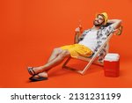 Small photo of Young smiling happy fun cool tourist man in beach shirt hat hold beer bottle lie on deckchair near fridge isolated on plain orange background studio portrait. Summer vacation sea rest sun tan concept