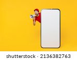 Full body delivery guy employee man in red cap T-shirt uniform workwear work as dealer courier stand behind mobile phone blank screen workspace shout in megaphone isolated on plain yellow background