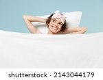 Young smiling happy woman in pajamas jam sleep eye mask rest relax at home lie in bed on pillow under blanket hold hand behind neck isolated on pastel blue background Good mood night bedtime concept.
