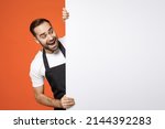 Small photo of Young excited man barista bartender barman employee in apron white t-shirt work coffee shop big white blank billboard for promo content text image place isolated on orange background Business startup