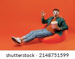 Full size body shoked fun young brunet man 20s wears red t-shirt green jacket sit in bag chairin 3d glasses watch movie film hold bucket of popcorn isolated on plain orange background studio portrait.