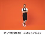 Full length young man barista bartender barman employee in black apron white t-shirt work in coffee shop hold hands crossed folded isolated on orange background studio. Small business startup concept
