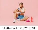 Small photo of Full size young sporty fitness trainer instructor woman wear blue tracksuit spend time in home gym sit on floor use mobile cell phone isolated on plain light pink background. Workout sport concept.