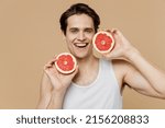 Small photo of Attractive satisfied fun young man he 20s perfect skin in undershirt hold in hand half of grapefruit isolated on plain pastel beige background studio. Skin care healthcare cosmetic procedures concept