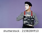 Small photo of Charismatic young mime man with white face mask wears striped shirt beret holding classic black film making clapperboard looking aside isolated on plain pastel light violet background studio portrait