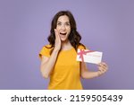 Surprised young brunette woman 20s wearing basic yellow t-shirt posing hold in hands gift certificate put hand on cheek looking camera isolated on pastel violet colour background, studio portrait