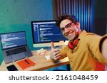 Small photo of Close up young software site reliability engineer IT programmer man in shirt work at home write code on laptop pc computer do selfie shot pov on mobile phone show thumb up Program development concept