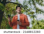 Small photo of Lower young man in orange jacket walk hold mobile cell phone takeaway delivery paper cup coffee to go rest relax in spring green city park go down alley sunshine lawn outdoor on nature. Urban concept