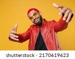 Young african cool rapper happy man 20s with funky trendy pink hairdo wear stylish red jacket glasses stretch hands to camera say yo yeah hi isolated on yellow orange color background studio portrait