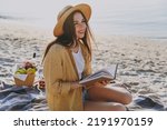 Full body young happy traveler tourist woman in straw hat shirt summer clothes reading book sit on plaid have picnic outdoors on sea sand beach background People vacation lifestyle journey concept.