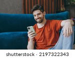 Small photo of Young man wears red t-shirt hold in hand use mobile cell phone sit on blue sofa couch stay at home hotel flat rest relax spend free spare time in living room indoors grey wall. People lounge concept