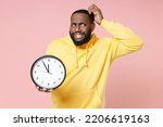 Small photo of Preoccupied young man of African American ethnicity 20s wearing casual yellow hoodie standing holding clock put hand on head looking aside isolated on pastel pink color wall background studio portrait