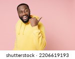 Small photo of Side view of funny young man of African American ethnicity 20s wear casual basic yellow hoodie stand pointing thumb aside on mock up copy space isolated on pastel pink color background studio portrait