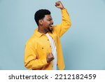 Small photo of Side view young fun man of African American ethnicity wear yellow shirt doing winner gesture celebrate clenching fists say yes isolated on plain pastel light blue background. People lifestyle concept