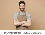 Young smiling happy man barista barman employee wear brown apron work in coffee shop hold hand crossed folded look camera isolated on plain pastel light beige background Small business startup concept