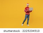 Full body merry young IT man wear red knitted Christmas sweater Santa hat posing look aside hold use work on laptop pc computer isolated on plain yellow background. Happy New Year 2023 holiday concept