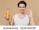 Small photo of Attractive young man 20s perfect skin in undershirt hold bottle of shampoo show ok gesture isolated on plain pastel beige background studio portrait. Skin care healthcare cosmetic procedures concept