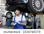 Young professional technician mechanic man wearing denim blue overalls t-shirt use hold mobile cell phone stand near car lift check technical condition work in vehicle repair shop workshop indoors.