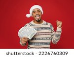 Merry excited cool young man wear Christmas sweater Santa hat posing hold fan of cash money in dollar banknotes do winner gesture isolated on plain red background. Happy New Year 2023 holiday concept