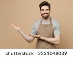 Young man barista barman employee wear brown apron work in coffee shop point hands arms aside indicate on workspace area isolated on plain pastel light beige background. Small business startup concept